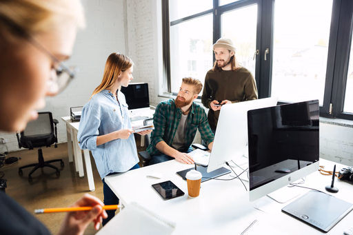 Coworkers discussing work around desktop monitors in a bright office.