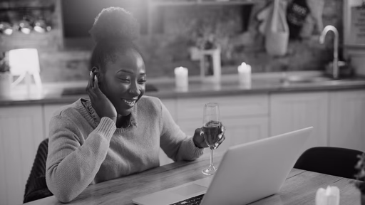 A woman on a phone call beside an open laptop at a kitchen table.