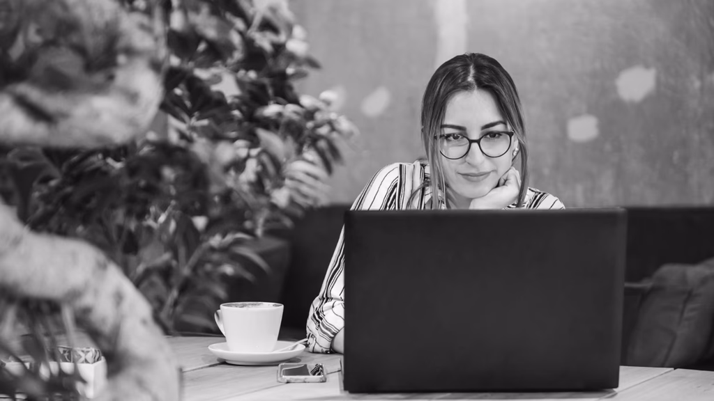A woman with glasses looking at a laptop beside a cup of coffee.